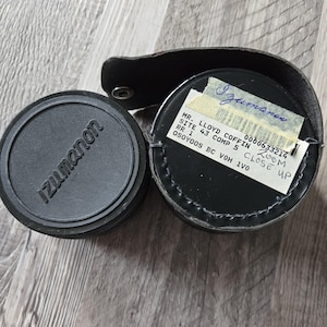 Puede incluir: Tapa de lente Izumanon negra junto a un estuche de cuero negro con una etiqueta que dice "MR. LLOYD COFFIN ZOOM SITE 43 COMP 5 CLOSE UP". El estuche está abierto, revelando la lente en su interior.