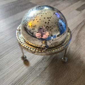 Puede incluir: Mantequillera plateada con tapa abovedada y detalles ornamentados. El plato se apoya en tres pequeños pies decorativos. La tapa presenta un motivo floral y un pequeño asa. El plato es de metal y tiene una superficie reflectante.