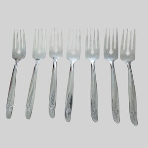 Könnte beinhalten: Ein Set aus sieben silberfarbenen Metallgabeln, die in einer Reihe angeordnet sind. Jede Gabel hat vier Zinken und einen Griff, der am unteren Ende mit einem erhabenen Blumenmotiv in Form einer Rose verziert ist. Das Metall ist glatt poliert.