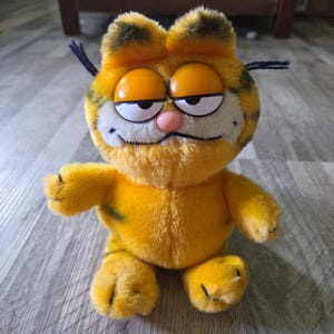 Könnte beinhalten: Ein Plüschtier in Form einer gelb-orangen getigerten Katze mit schwarzen Streifen, großen Kunststoffaugen mit halb geschlossenen Lidern, einer kleinen rosa Nase und schwarzen Schnurrhaaren. Die Figur zeigt ein breites, dezentes Lächeln.