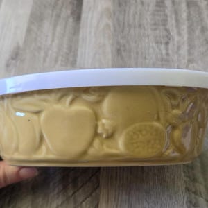 Puede incluir: Un cuenco redondo de cerámica de color tostado con un patrón de frutas en relieve en el exterior, que incluye representaciones de manzanas y granadas. El cuenco está cubierto por una tapa circular de plástico blanco que encaja.