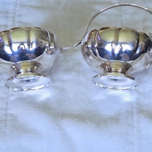 Op de afbeelding: Een paar zilverkleurige serveerstukken, één met een handvat en tuit, de andere met twee kleine handvatten. Beide hebben een afgerond, geribbeld lichaam en staan op een voetstuk. De reflecterende oppervlakken tonen de omgeving.