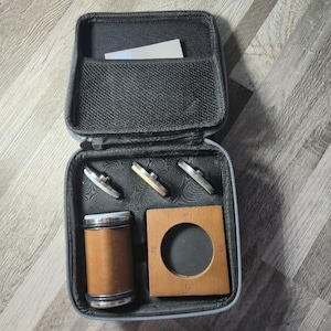 May include: An open black zippered case containing various tools. The case holds three silver and gold-toned metal objects, a wooden square with a circular cutout, and a cylindrical brown and silver object. A small white card is in the top pocket.