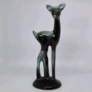 Puede incluir: Una figura de ciervo de cerámica de pie con un acabado negro brillante y acentos moteados en verde azulado en las orejas, el lomo y las patas. La escultura tiene una superficie lisa y reflectante sobre una base circular.