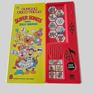 May include: A Golden Sing Along Super Songs with Silly Sounds book and a red plastic electronic sound player. The yellow book cover features cartoon characters and the text 8 Songs to Play and Sing. The red player has buttons with icons.