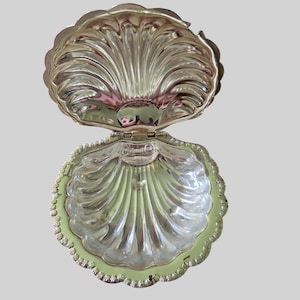 May include: A silver-toned metal clam shell dish with a hinged lid. The interior and exterior surfaces feature a ribbed, scalloped design with a polished, reflective finish. The object is open, revealing the concave bottom and the lid.