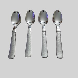 May include: A set of four silver-toned metal spoons arranged in a row. Each spoon features a smooth, polished oval bowl and a textured, hammered-finish handle that provides a distinct contrast to the reflective surface of the spoon head.