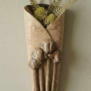 May include: A wall-mounted ceramic pocket planter with a speckled beige finish, featuring a sculpted cluster of three brown mushrooms on the front. The vessel contains green fern fronds and light green reindeer moss, suspended by a thin cord.