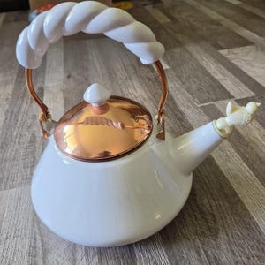 May include: A white tea kettle with a glossy finish, featuring a copper-coloured lid and a white twisted rope-style handle. The spout is topped with a small, cream-coloured bird figurine. The kettle has a rounded base and a matching white lid knob.