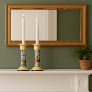 Op de afbeelding: Twee brandende witte kaarsen in decoratieve gele en bloemmotief kandelaars. De kandelaars hebben een blauw, oranje en geel bloemmotief. Een goudomrand spiegel hangt op de achtergrond.