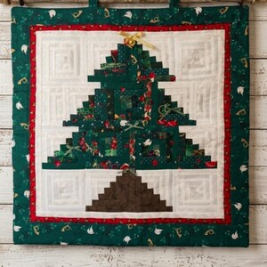 May include: A quilted Christmas tree wall hanging. The tree is made of green fabric with festive patterns, and is set against a white background with a red border. The outer border is green with white and gold accents. A gold bow adorns the top.