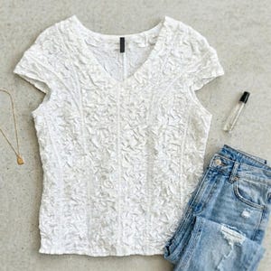 Può includere: Un top bianco strutturato con scollo a V e maniche corte, abbinato a jeans di denim blu chiaro. Sono visibili anche una collana d'oro con un piccolo ciondolo e un flacone di profumo. Gli articoli sono disposti su una superficie grigia.