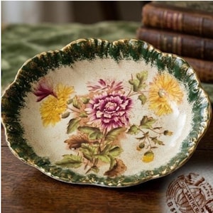 May include: An oval ceramic dish with a cream-colored base featuring a crackled glaze finish. The center displays a floral illustration of purple and yellow chrysanthemums with green leaves. The scalloped rim is dark green with gold trim.