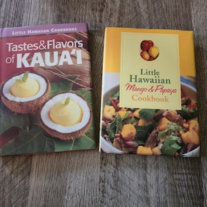 May include: Two cookbooks. One is titled "Tastes & Flavours of Kauai" and the other is "Little Hawaiian Mango & Papaya Cookbook." The first book shows coconut shells filled with yellow food. The second book has a picture of a salad.