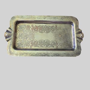 Puede incluir: Una bandeja de servicio rectangular de metal plateado con un patrón de volutas en relieve ornamentado en el borde ancho y el área central empotrada. La bandeja incluye dos asas integradas en los lados cortos con detalles decorativos.
