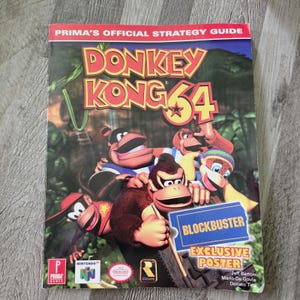 May include: A Prima's Official Strategy Guide for Donkey Kong 64. The cover features Donkey Kong and other characters from the game in a jungle setting. A blue Blockbuster Exclusive Poster sticker is on the front, along with Nintendo 64 branding.