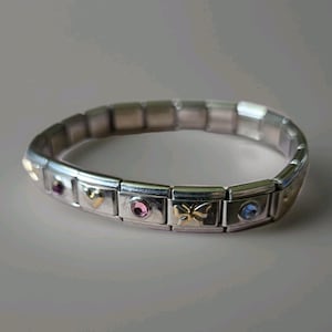 Könnte beinhalten: Ein modulares Gliederarmband in Silberoptik mit abwechselnden rechteckigen Gliedern. Einige Glieder sind schlicht, während andere mit einem goldfarbenen Herz, einem goldfarbenen Schmetterling und kleinen runden Edelsteinen in Rosa und Hellblau verziert sind.