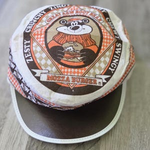 Könnte beinhalten: Eine weiß-orange gemusterte Kappe mit einer zentralen Grafik eines Bären, der einen Burger hält. Der Text lautet Great A&W Root Bear, Zesty, Crunchy, Zingy, Chewy, Swingy und Mozza Burger. Die Kappe hat einen dunkelbraunen Schirm mit weißem Rand.