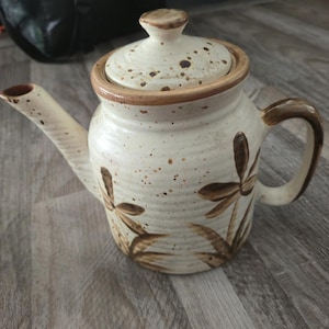 Pode incluir: Bule de cerâmica creme com tampa, bico e alça. O bule apresenta desenhos florais castanhos e detalhes salpicados. A tampa e a borda têm uma borda castanha. O bule está sobre uma superfície de madeira.
