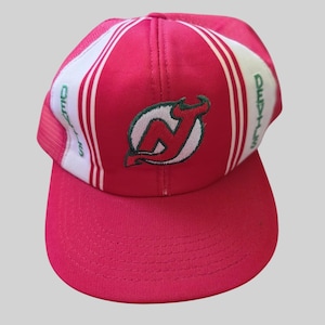 May include: A bright pink and white baseball cap featuring the New Jersey Devils logo embroidered in red, white, and green on the front panel. The word Devils is embroidered in green on the side panels, and the cap has a flat brim.