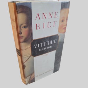以下が含まれることがあります： アン・ライス著『Vittorio the Vampire』のハードカバー本。ダストジャケットには、色白でダークヘアの人物の分割された肖像画が描かれており、白い背景に赤い文字で著者名、黒い文字でタイトルが記されています。