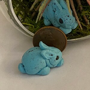 May include: Two small blue ceramic bunnies with speckled paint. The bunnies are lying down and are about the size of a UK penny.