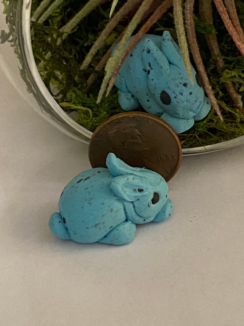 May include: Two small blue ceramic bunnies with speckled paint. The bunnies are lying down and are about the size of a UK penny.
