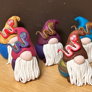 May include: Six miniature gnomes with white beards and colorful hats. Each gnome has a small snake on their hat. The snakes are different colors, including red, purple, and brown.