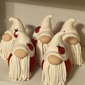 May include: Five white gnomes with red hats and white beards. Each gnome has a ladybug on their hat. The gnomes are sitting on a white surface.