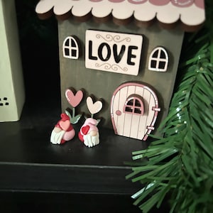 May include: A wooden house-shaped decoration with a pink roof and the word "LOVE" on the front. Two small gnomes with heart-shaped accents and a pink door are also featured. The house is decorated with pink and white accents.
