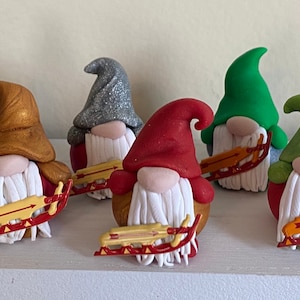 May include: Seven handmade gnome figurines with white beards and red hats. Each gnome is holding a small wooden sled with a yellow arrow painted on it. The gnomes are made of polymer clay and have different colored hats, including green, gold, silver, and red.