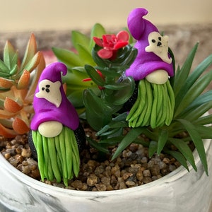 May include: Two purple and green Halloween gnomes with ghost faces on their hats are sitting in a white pot with green and red plants.