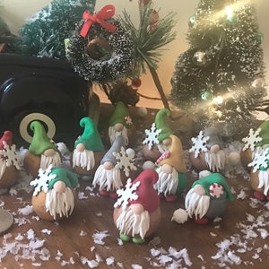 May include: A collection of 14 miniature Christmas gnomes made of polymer clay. Each gnome is wearing a different colored hat and has a white beard. Some gnomes are wearing a snowflake, candy cane, or a yellow and red striped hat.