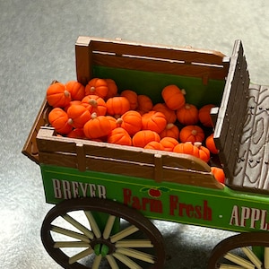 May include: A green toy wagon with wooden wheels and a wooden bed filled with orange pumpkins. The wagon has the words "Breyer Farm Fresh Apples" printed on the side.