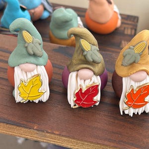 May include: Three ceramic gnome figurines with autumn-themed details. Each gnome has a long white beard, a colourful leaf, and a hat with a corn cob. The gnomes are on a wooden surface.