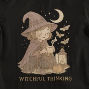 May include: Black t-shirt featuring a whimsical illustration of a person in a witch hat, cape, and holding a cup, with a cat, lantern, and butterflies. The text "WITCHFUL THINKING" is printed below the illustration.