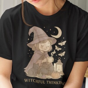 May include: Black t-shirt with a whimsical illustration of a witch with a hat, cape, and a cup of tea, surrounded by butterflies and a crescent moon. The text "WITCHFUL THINKING" is below the illustration.