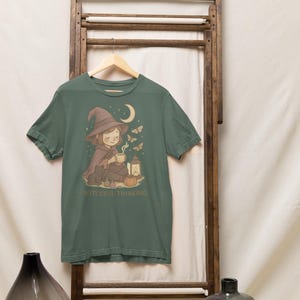 May include: Green t-shirt featuring a whimsical illustration of a witch with a hat, cape, and a cup, with a moon, butterflies, and a lantern. The text "WITCHFUL THINKING" is printed below the illustration.