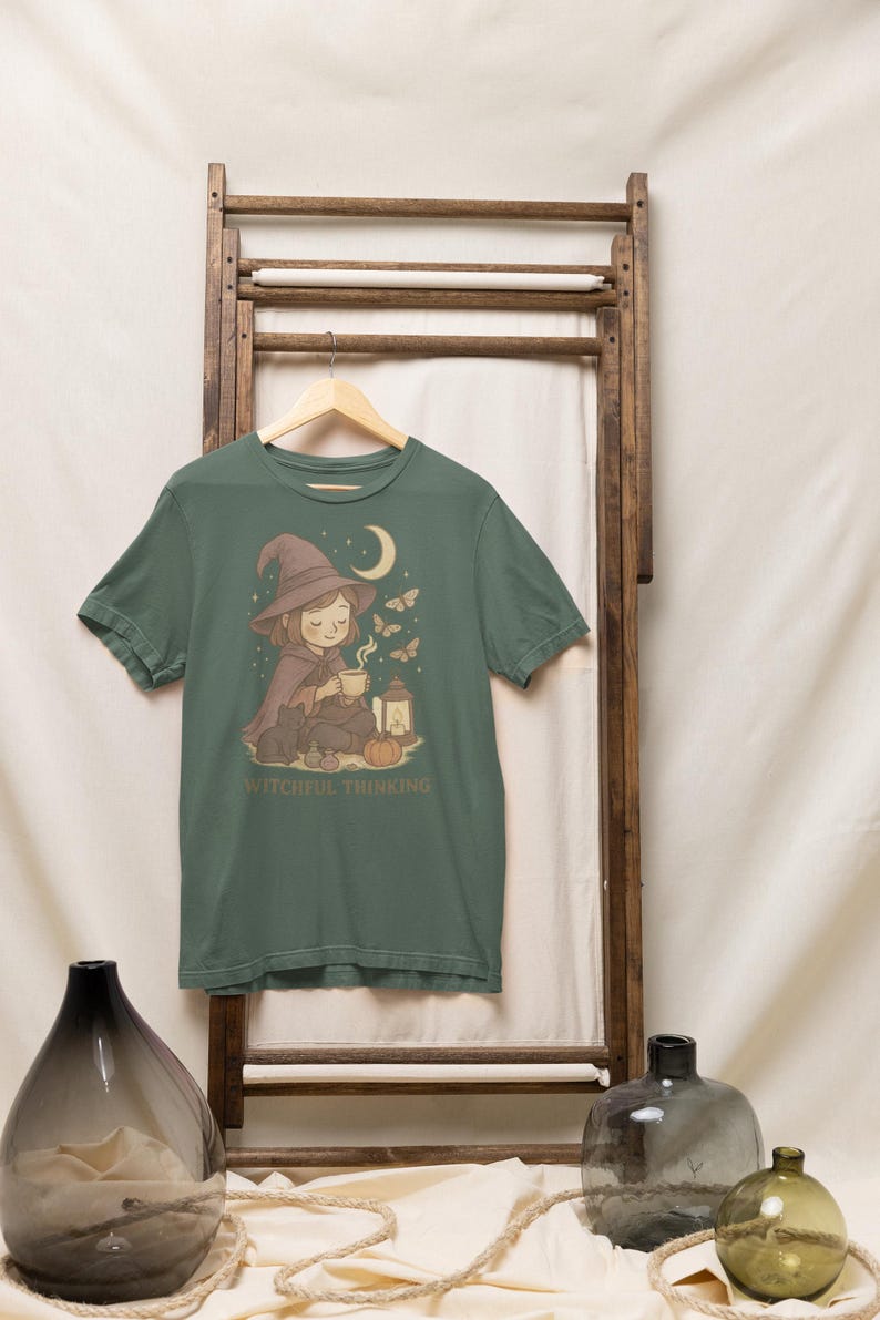 May include: Green t-shirt featuring a whimsical illustration of a witch with a hat, cape, and a cup, with a moon, butterflies, and a lantern. The text "WITCHFUL THINKING" is printed below the illustration.