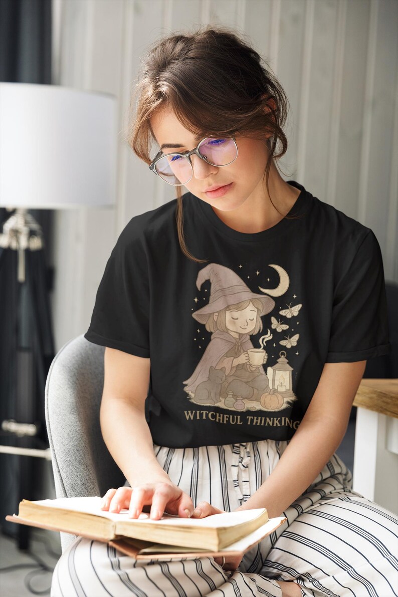 May include: Black t-shirt with a whimsical illustration of a witch with a hat, cape, and a cup of tea, surrounded by butterflies and a crescent moon. The text "WITCHFUL THINKING" is below the illustration.