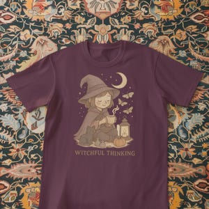 May include: A maroon t-shirt featuring a whimsical illustration. It depicts a seated figure in a witch's hat and cape, holding a cup, with a black cat, a lantern, and the text "Witchful Thinking". Butterflies and a crescent moon are also present.