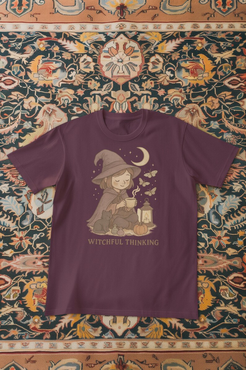 May include: A maroon t-shirt featuring a whimsical illustration. It depicts a seated figure in a witch's hat and cape, holding a cup, with a black cat, a lantern, and the text "Witchful Thinking". Butterflies and a crescent moon are also present.