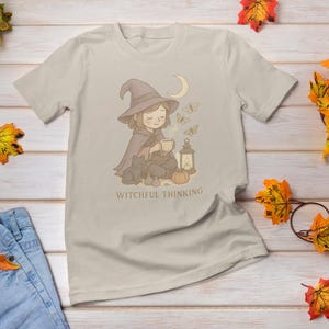 May include: Beige t-shirt featuring a whimsical illustration of a witch with a hat, cape, and a cat, holding a cup. The design includes butterflies, a moon, and a lantern. Text reads "Witchful Thinking".