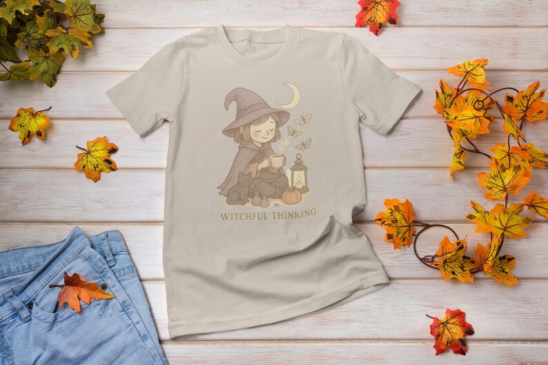 May include: Beige t-shirt featuring a whimsical illustration of a witch with a hat, cape, and a cat, holding a cup. The design includes butterflies, a moon, and a lantern. Text reads "Witchful Thinking".