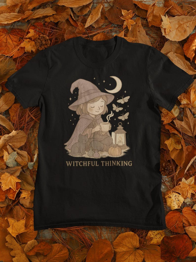 May include: Black t-shirt featuring a whimsical illustration of a person in a witch hat, cape, and holding a cup, with a cat, lantern, and butterflies. The text "WITCHFUL THINKING" is printed below the illustration.