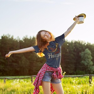 May include: A navy blue t-shirt with the word "Huzzah" in a decorative font, along with a horn graphic. The person in the image is wearing a red and black plaid shirt and denim shorts, holding a hat.