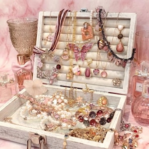 May include: A white jewellery box overflowing with necklaces, bracelets, and earrings in pink, gold, and pearl tones. A butterfly pendant and perfume bottles add to the feminine theme. The box is surrounded by glassware.