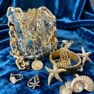 Op de afbeelding: Een verzameling gouden en zilveren sieraden, waaronder kettingen, armbanden, ringen en bedels, gepresenteerd op een blauw fluwelen oppervlak. De sieraden hebben schelp-, zeester- en vlindermotieven. Een glazen pot is gevuld met diverse kettingen en hangers.