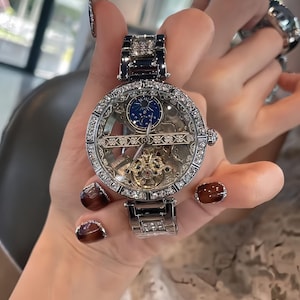 Könnte beinhalten: Eine silberfarbene Armbanduhr mit transparentem Zifferblatt, die das Innenleben zeigt. Die Uhr hat eine diamantbesetzte Lünette und ein Armband mit einem kleinen blauen Zifferblatt. Die Uhr wird von einer Person gehalten.