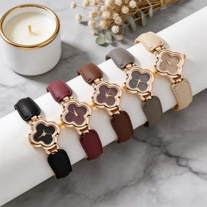 Puede incluir: Cinco relojes dorados con esferas en forma de trébol y correas de cuero en varios colores: negro, burdeos, marrón, gris y beige. Las esferas tienen un diseño sencillo. Una vela blanca en un soporte dorado está en el fondo.